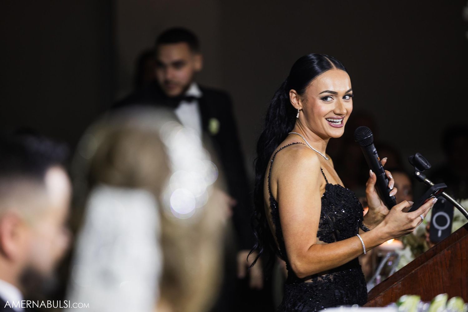 A woman in an elegant black dress speaks at a wedding reception podium.