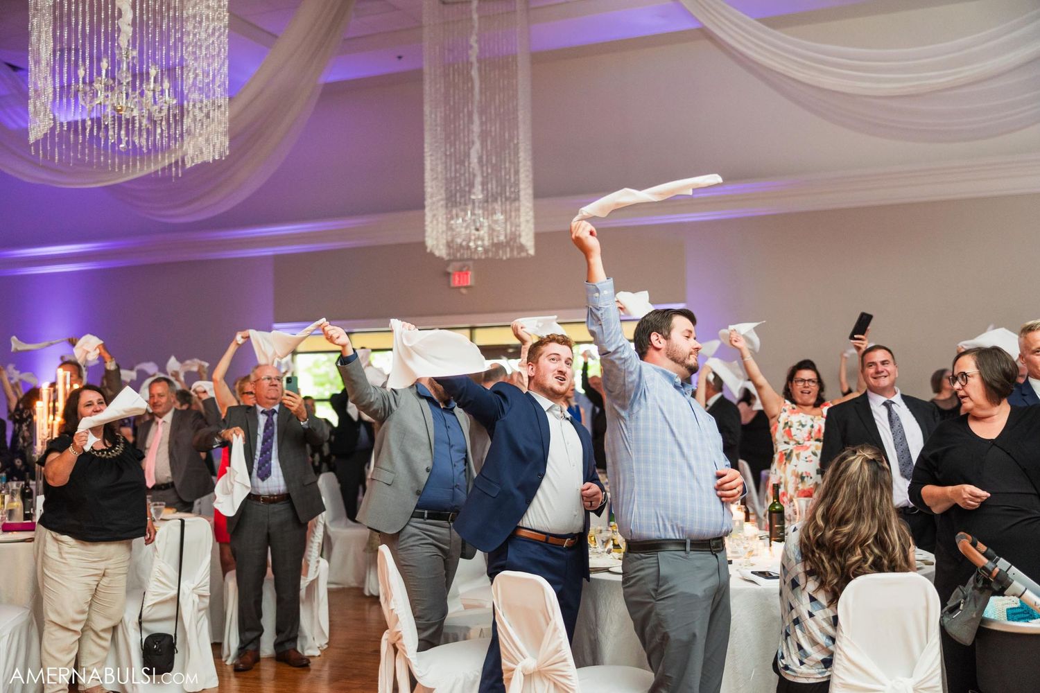 Wedding celebration with guests waving napkins under crystal chandeliers and white draping.