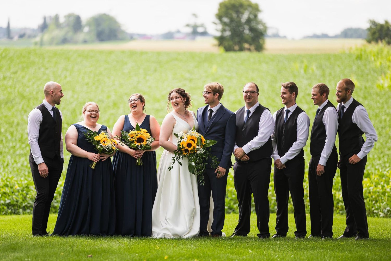 Wedding party in black attire poses together in a sunny green field with vibrant sunflowers.