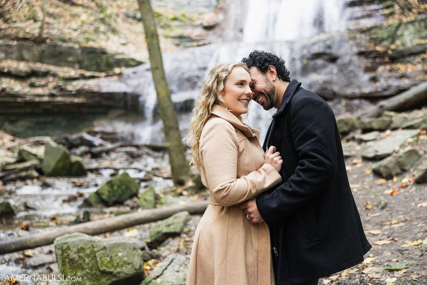 A couple in warm clothing shares a tender moment in front of a forest waterfall.