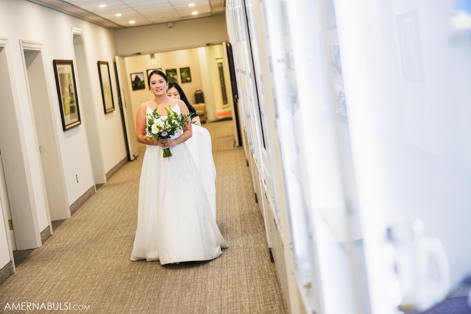 Bride walks down hallway in flowing white wedding gown holding elegant bouquet.