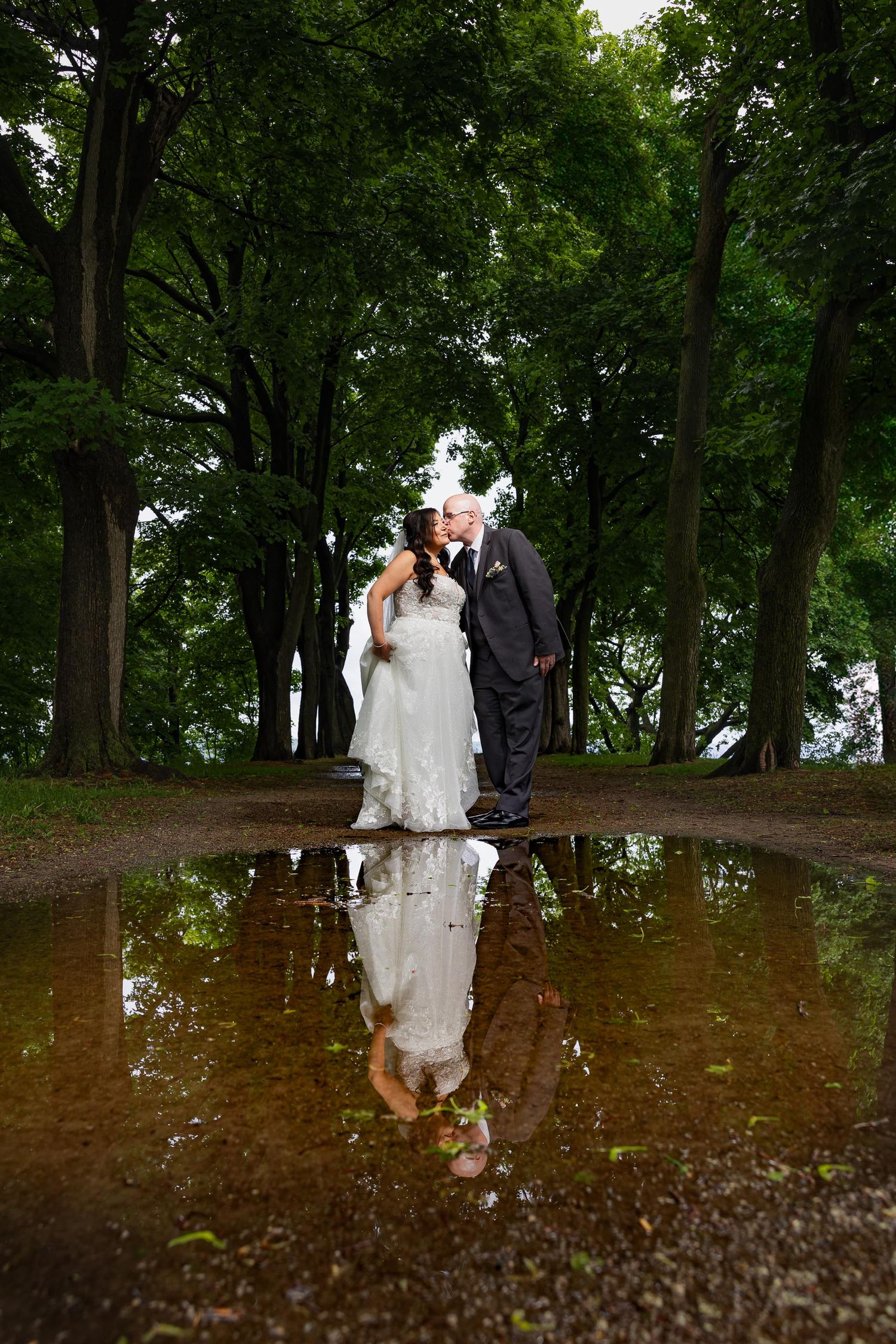 A couple shares a romantic moment with their reflection in a puddle surrounded by trees.