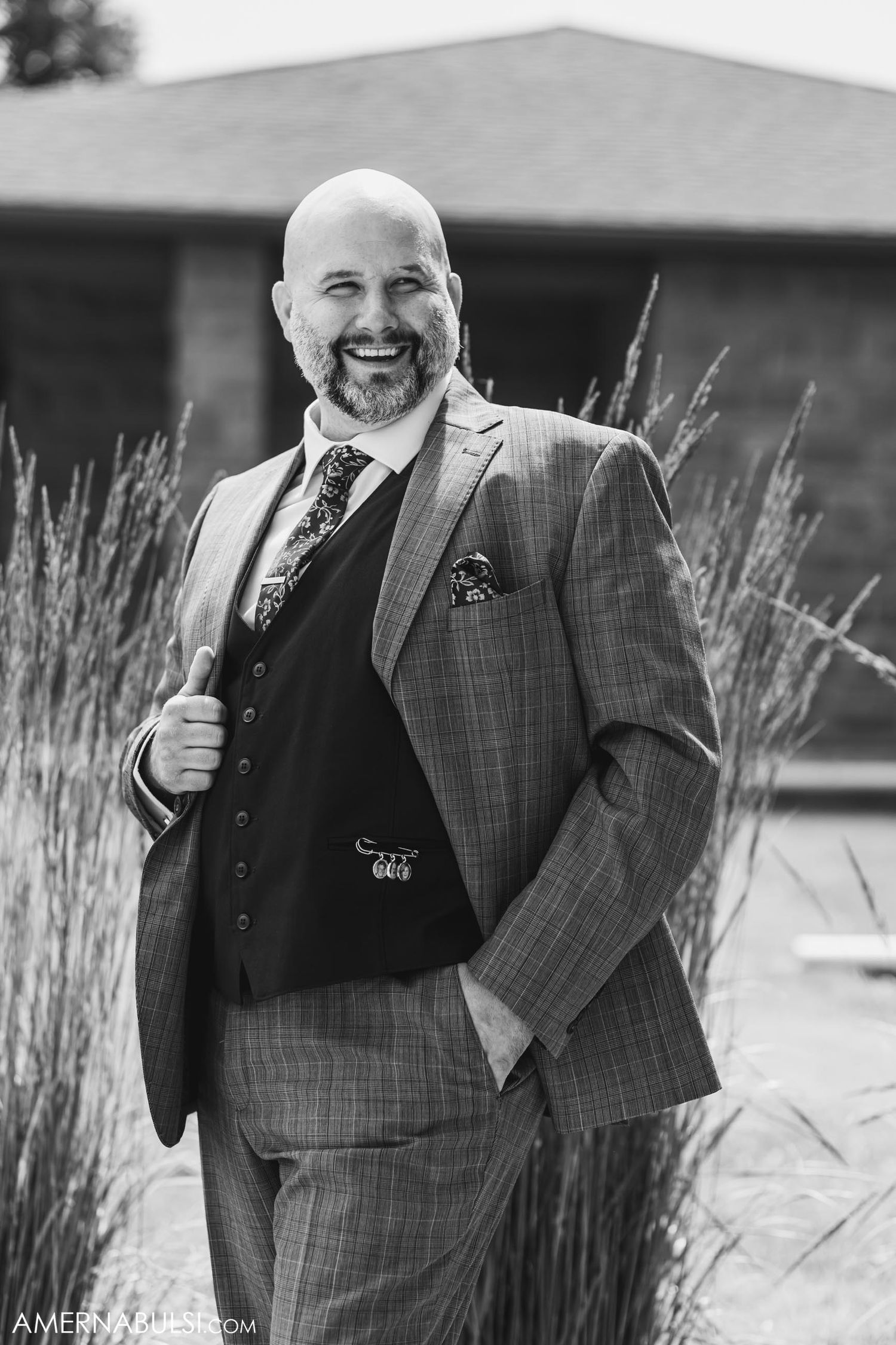 Man in gray plaid suit and vest poses outdoors near ornamental grasses.