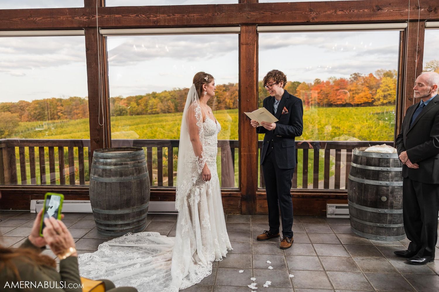 A wedding ceremony takes place on a rustic porch with wooden barrels and autumn foliage in the background.