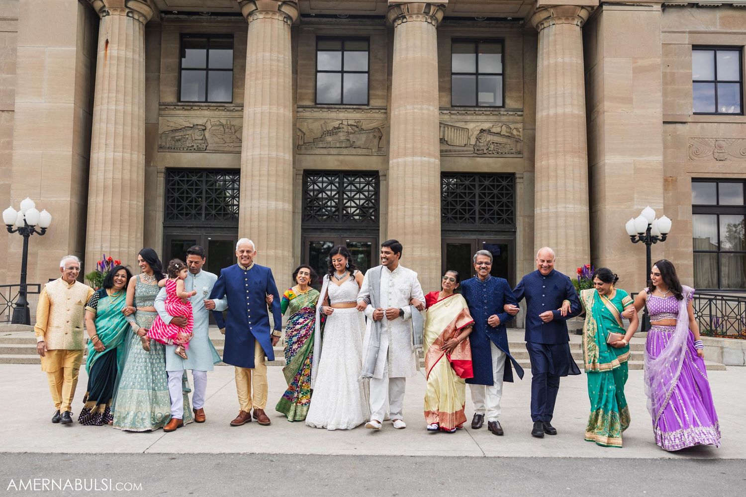 Wedding party members in colorful traditional Indian and Western attire pose on grand building steps.