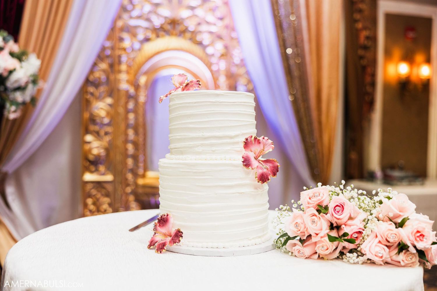 White tiered wedding cake decorated with pink flowers on display at elegant reception.