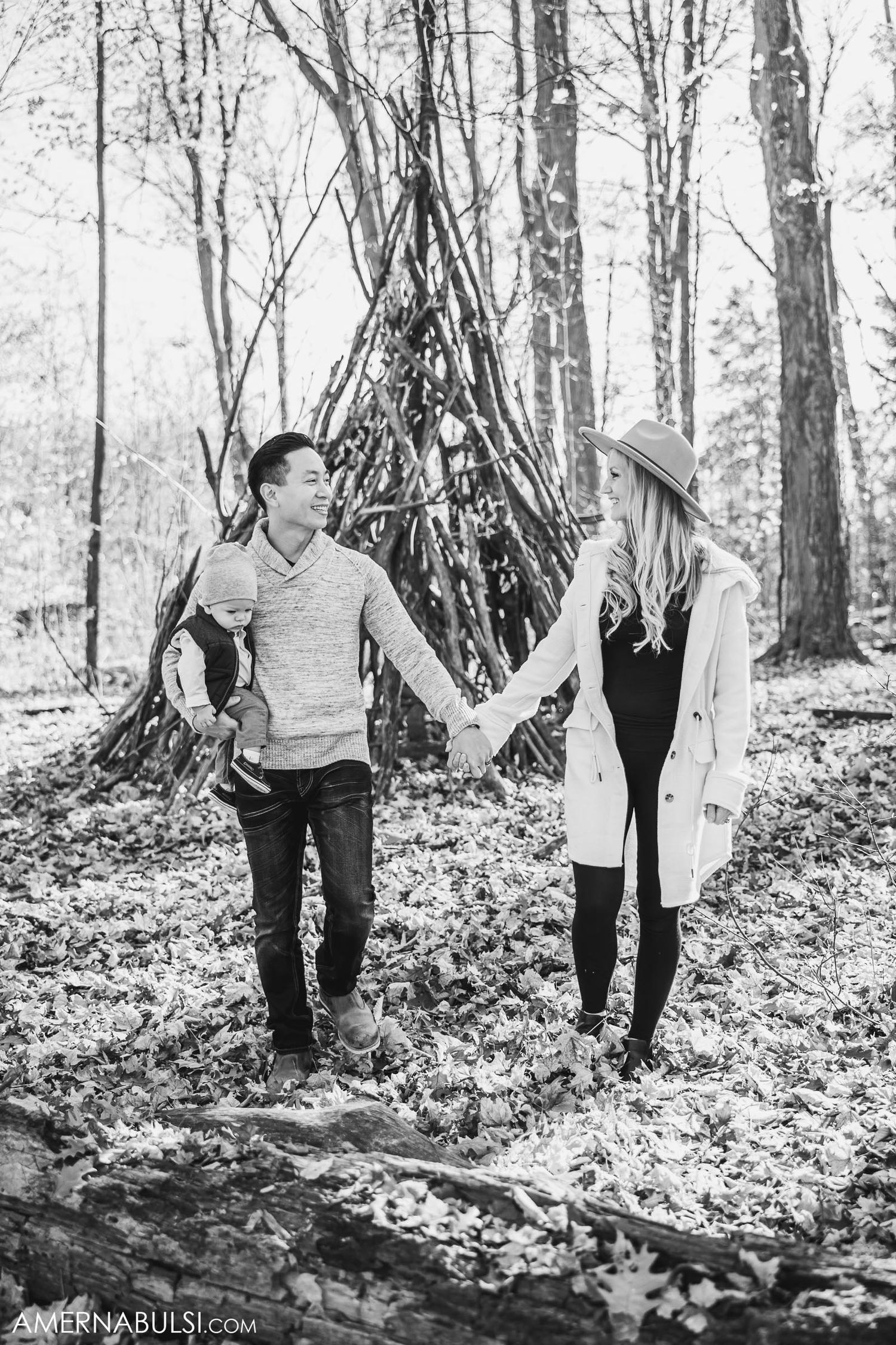 Black and white photo of couple holding hands by rustic teepee structure in woods.