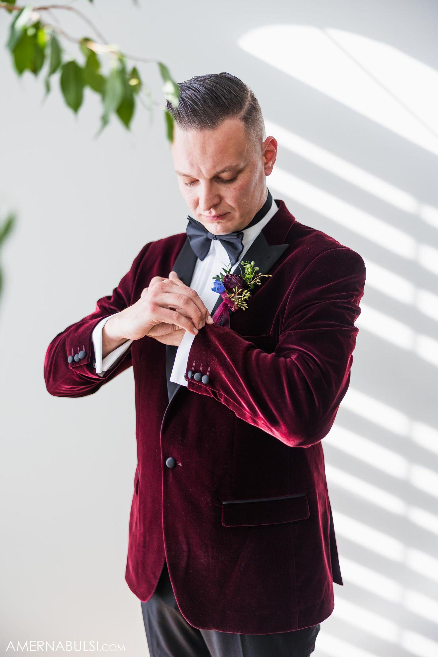 Groom in burgundy velvet jacket adjusts boutonniere near window with dramatic lighting.