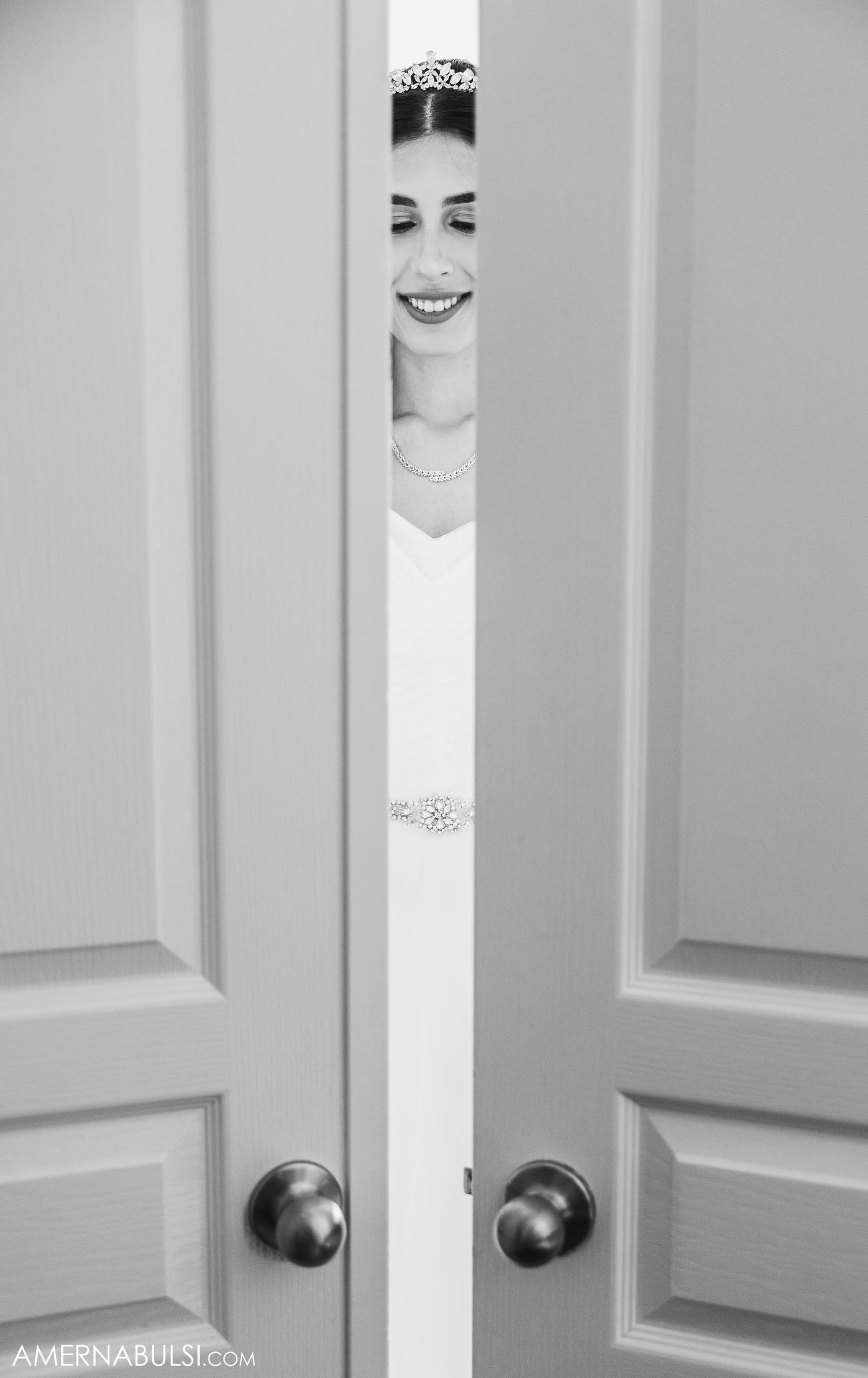 A smiling bride peeks through a small gap between elegant white doors wearing a sparkling tiara.