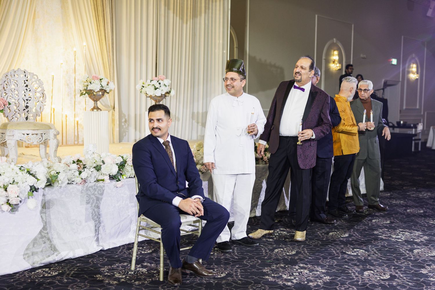 A group stands near an elegantly decorated head table at a formal event with white and gold decor.