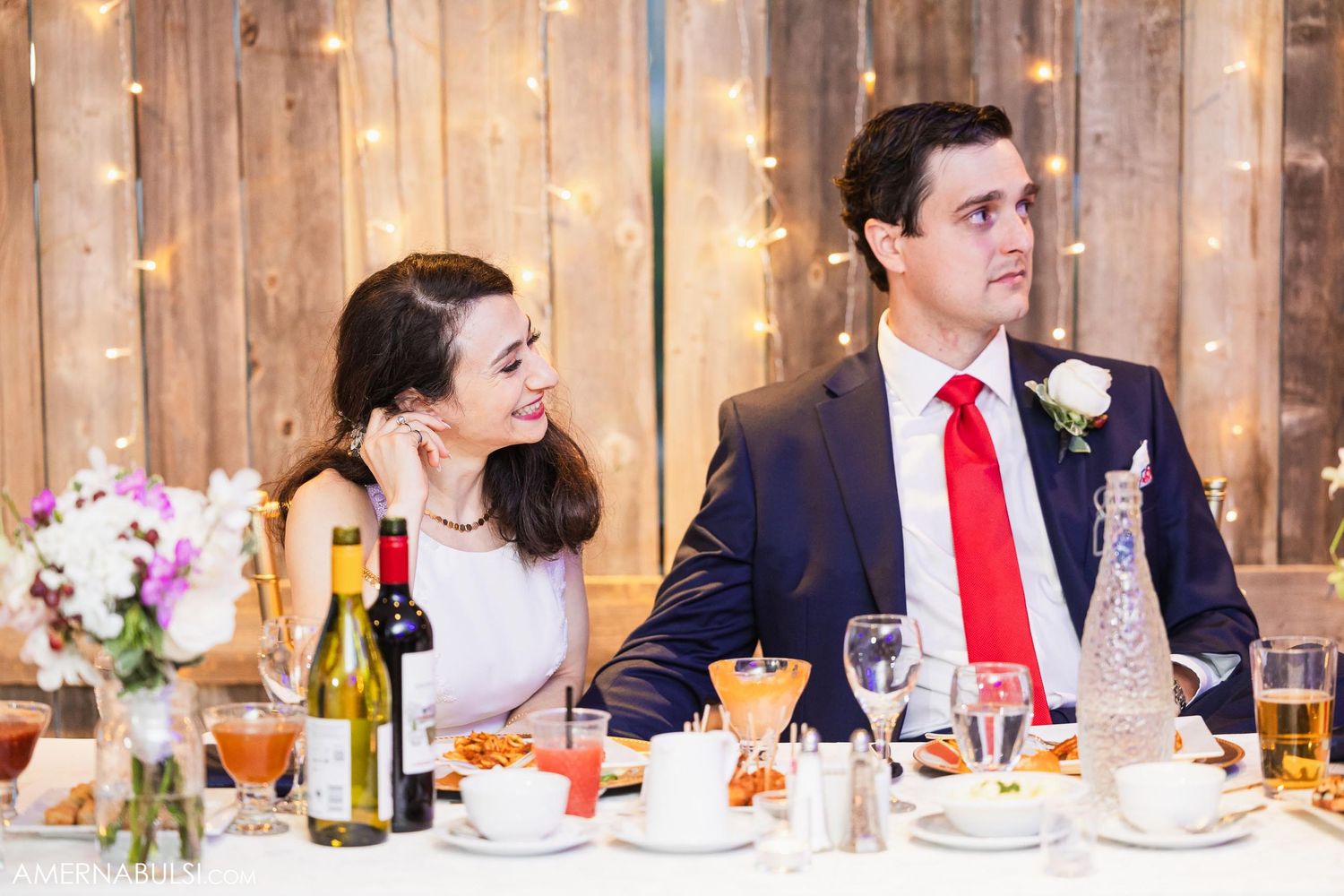 Wedding couple enjoying dinner at rustic wood paneled reception with string lights and floral centerpieces.