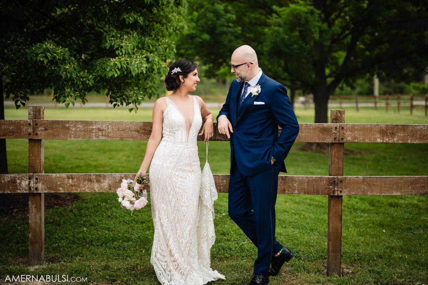 Couple in wedding attire walks together along wooden fence in outdoor garden setting.