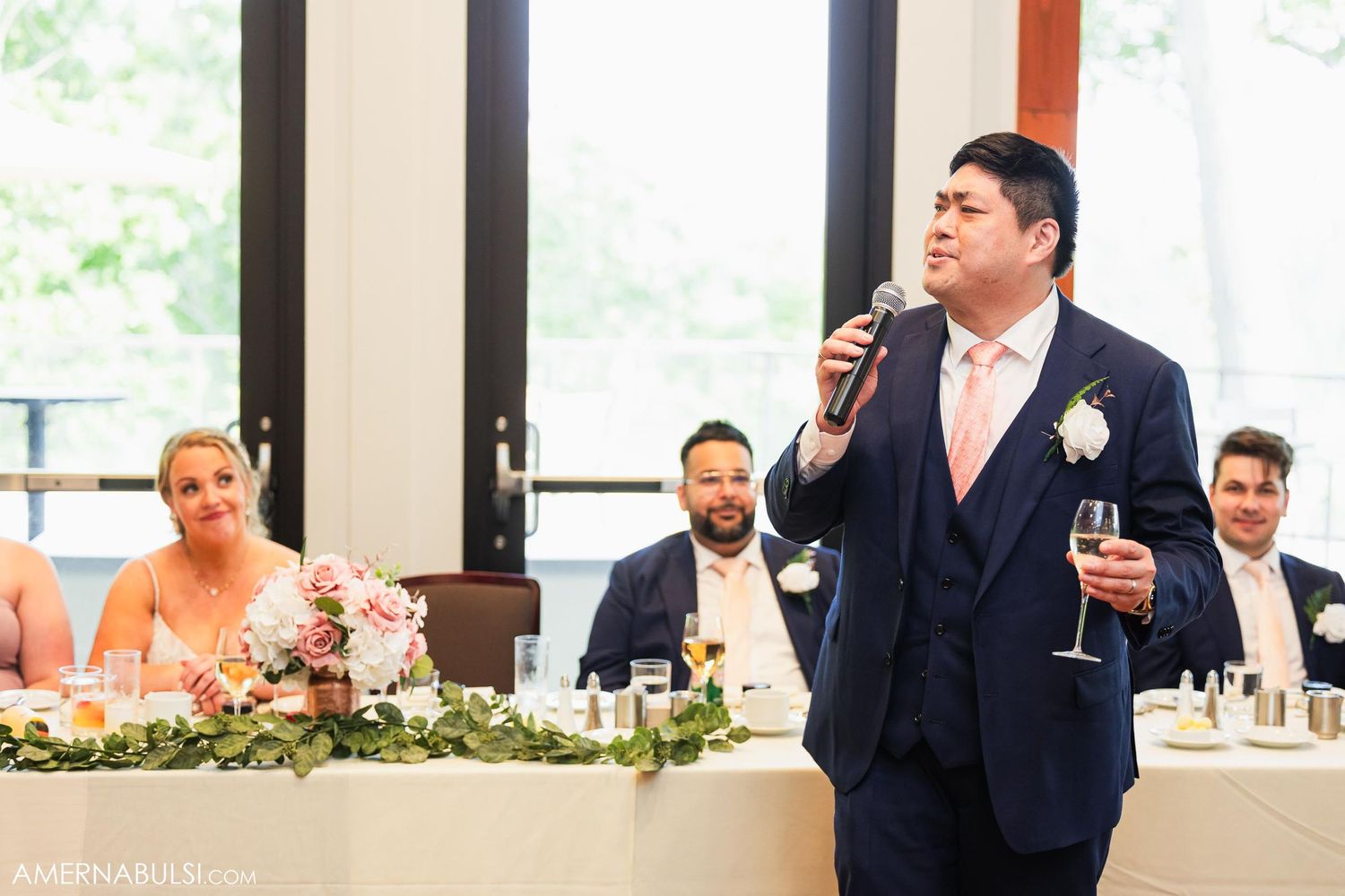 Wedding guest in navy suit delivers heartfelt speech at elegant reception with floral centerpiece decor.