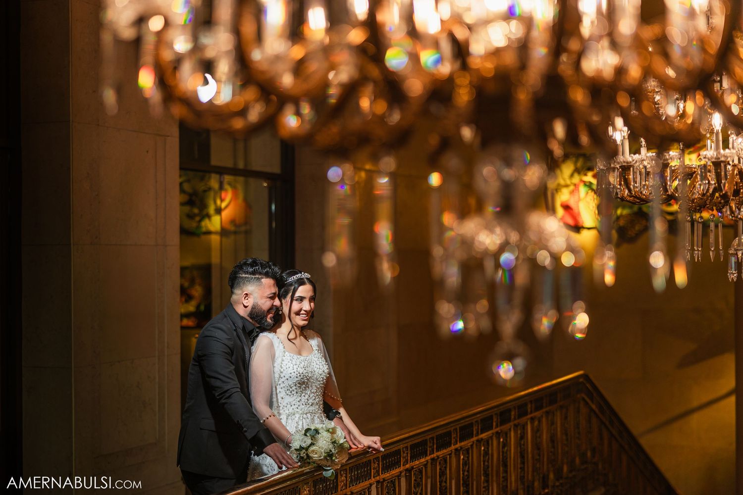 A newlywed couple stands on an elegant staircase under glowing crystal chandeliers.