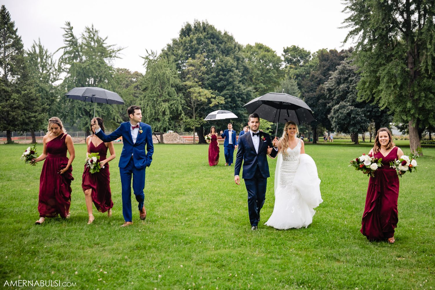 Wedding party walking with umbrellas across a rainy green lawn.