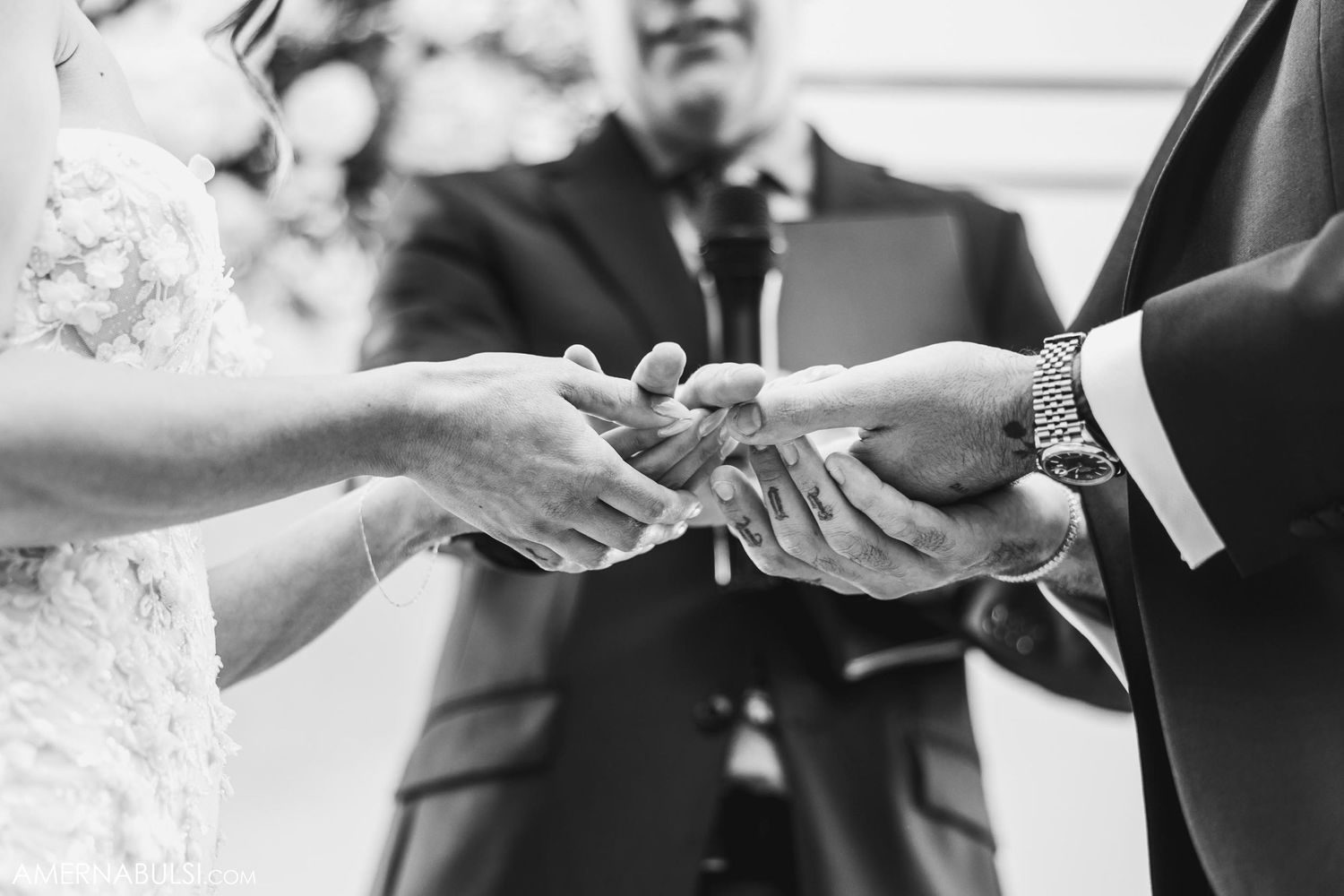 Black and white close-up of exchanging wedding rings during ceremony with officiant in background.
