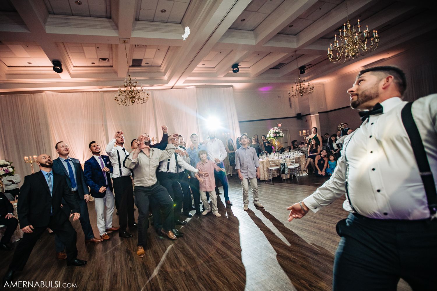 Wedding guests dance and celebrate on a wooden ballroom floor under elegant chandeliers at an evening reception.