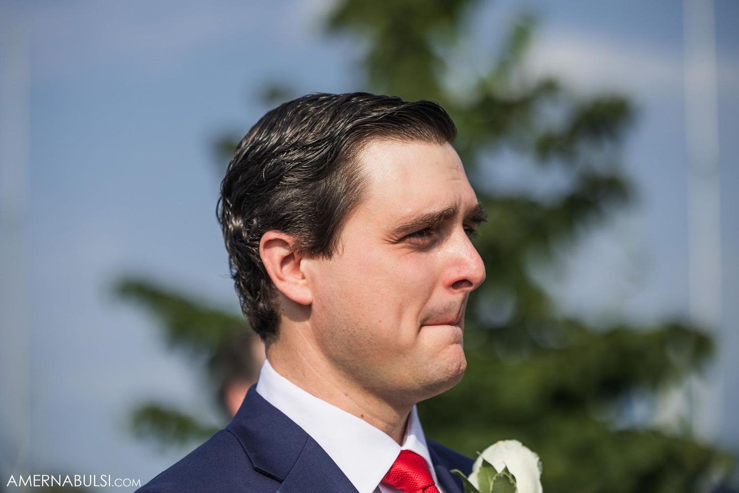 A groom wearing a navy suit with red tie and white boutonniere in an outdoor setting.