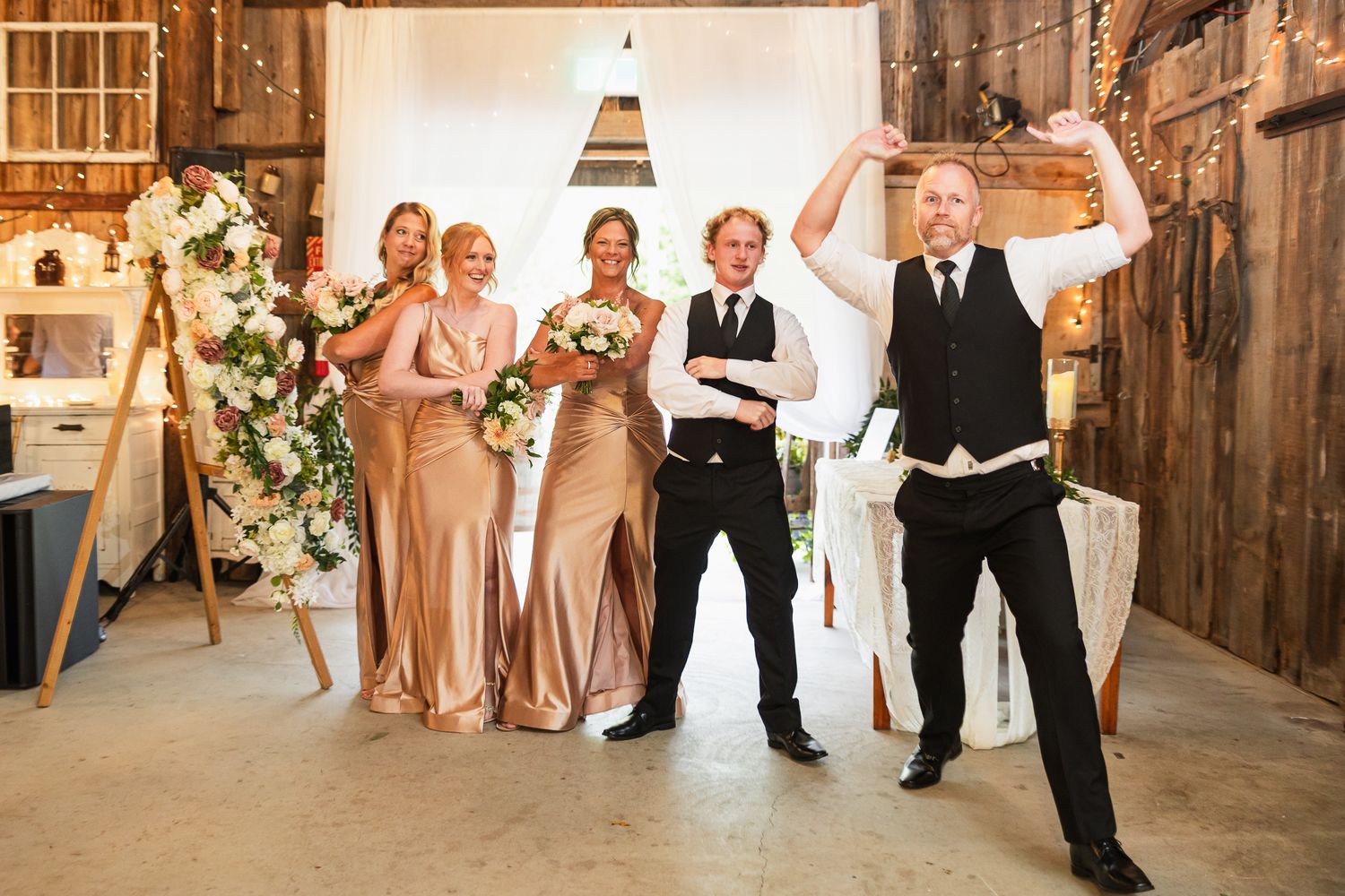 Wedding party posing in a rustic barn venue with bridesmaids in champagne dresses and groomsmen in black suits.