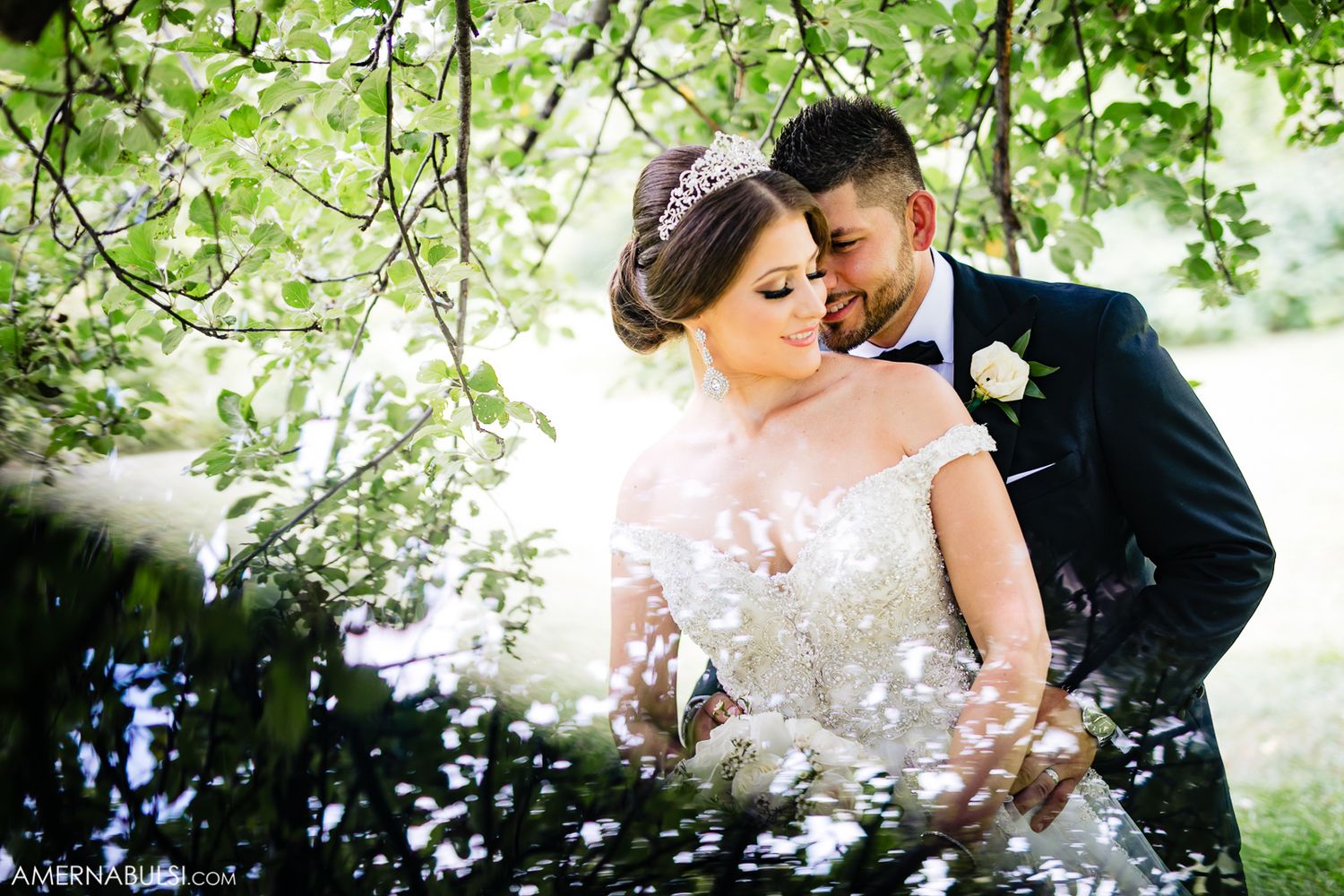 Wedding couple poses under lush green trees with sunlight filtering through the leaves.