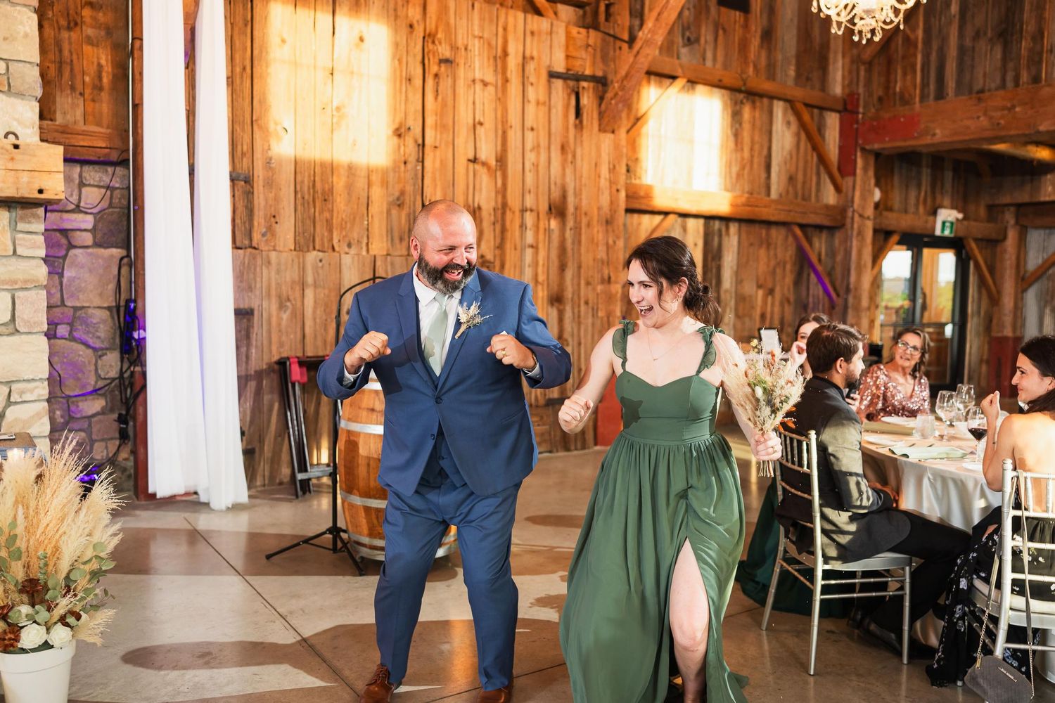 Wedding party members dance with enthusiasm in rustic barn reception venue.