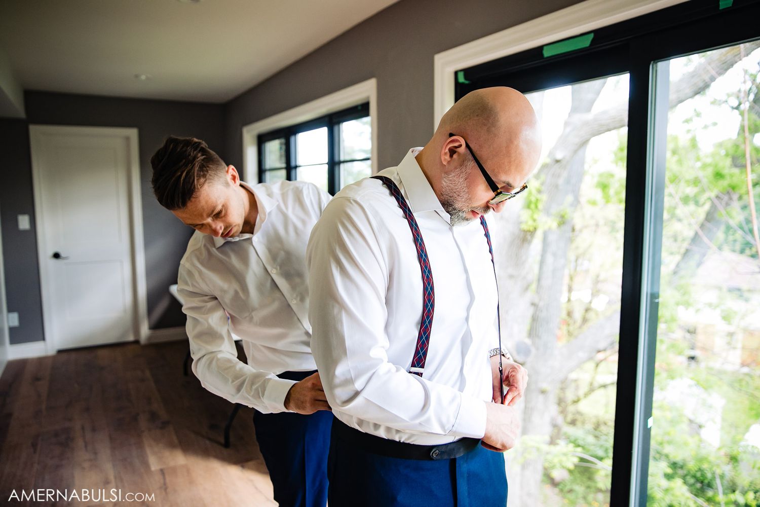 Two people in formal attire getting ready in a bright room with large windows.