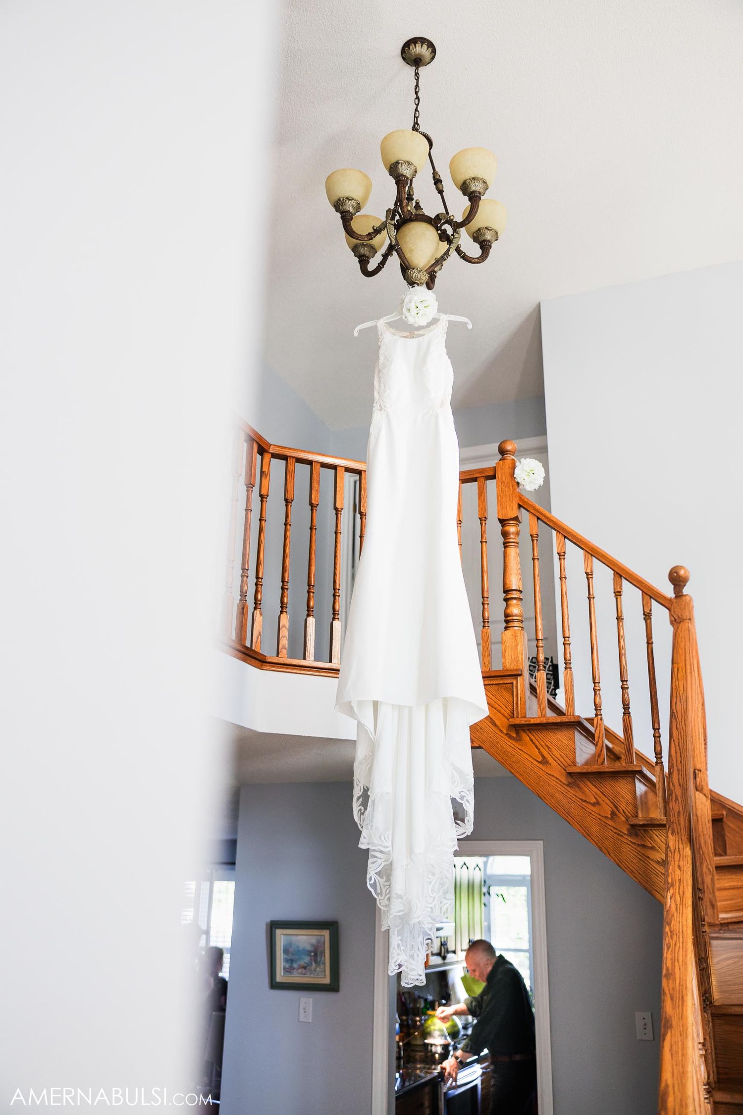 White wedding dress hanging from chandelier above wooden staircase in home interior.