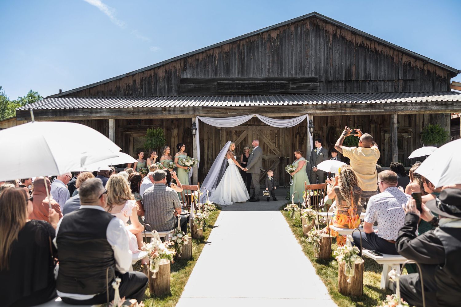 Outdoor rustic barn wedding ceremony with guests seated along white aisle under sunny blue sky.