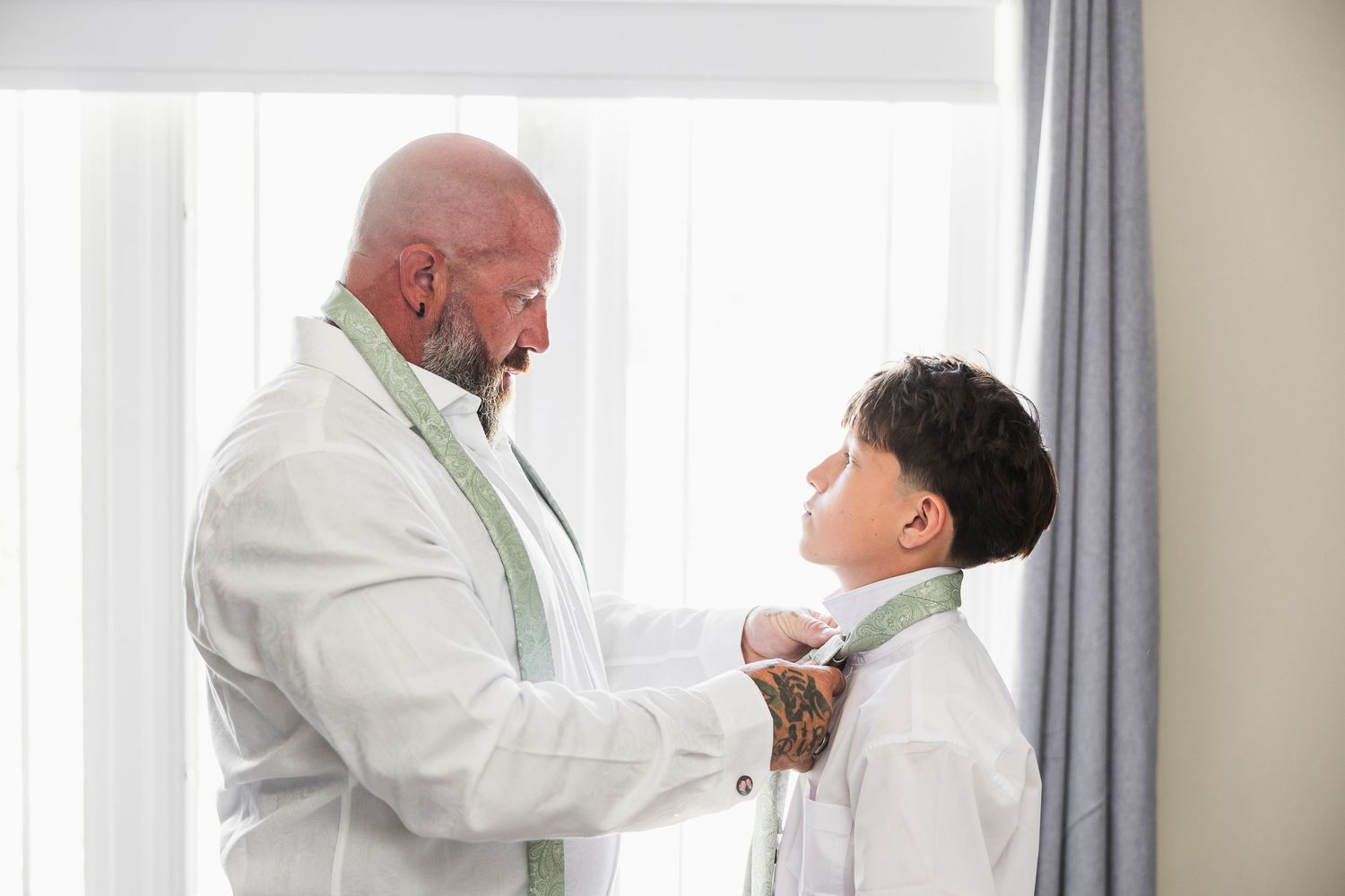 A father helps his young son adjust his tie, both dressed in matching white suits for a formal occasion.