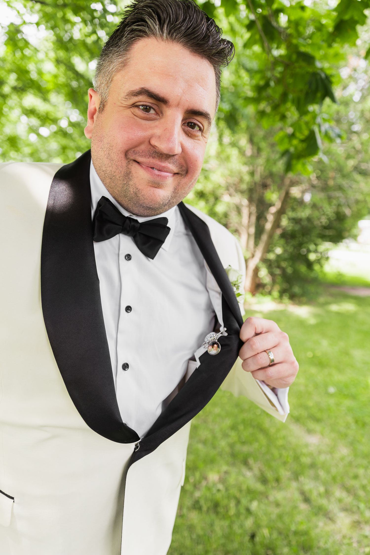 Smiling groom in white tuxedo with black bow tie adjusting boutonniere outdoors.