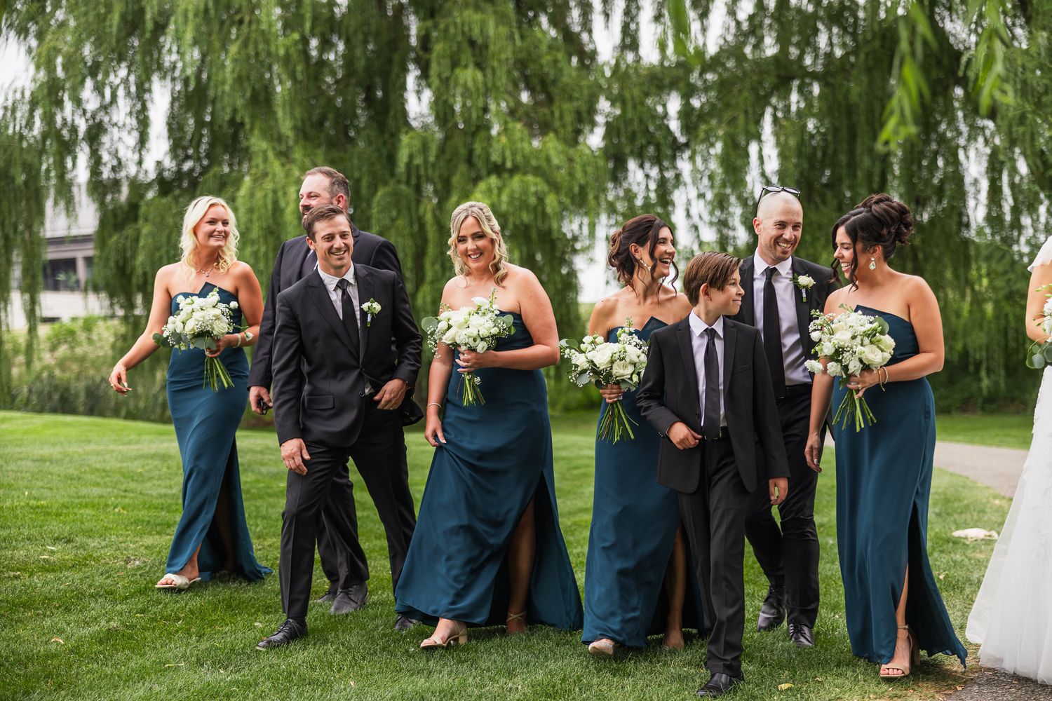 Wedding party in teal gowns and black suits walk together outdoors near weeping willow trees.