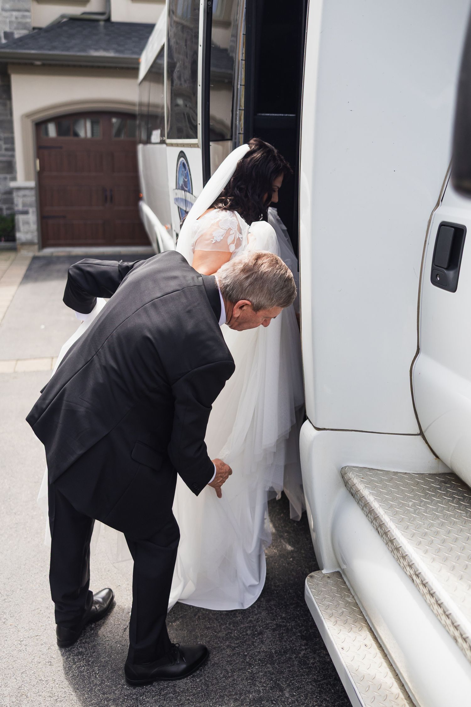 Father helping bride in white gown board white limousine bus.