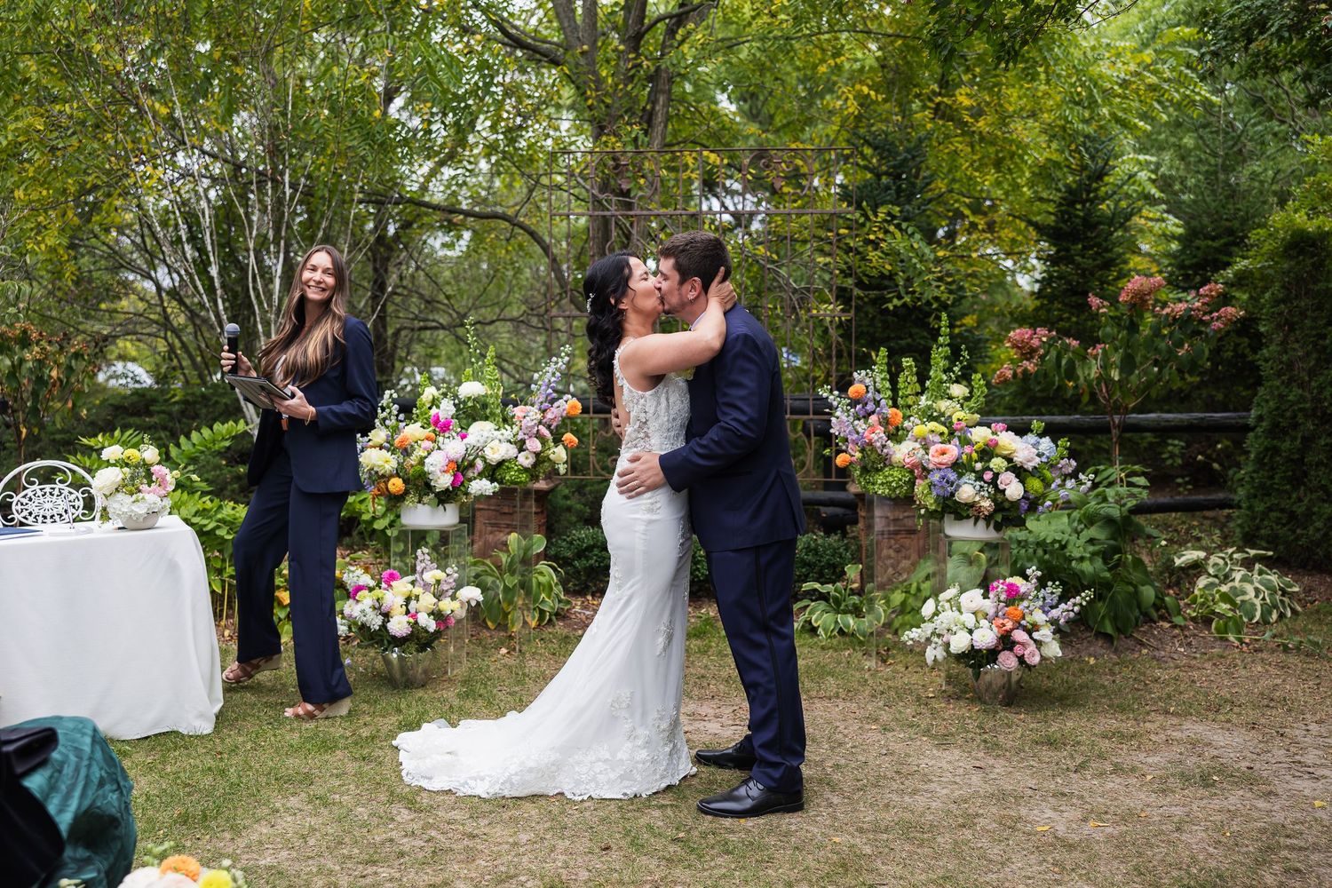 Bride and groom share their first kiss at an outdoor wedding ceremony surrounded by colorful floral arrangements.
