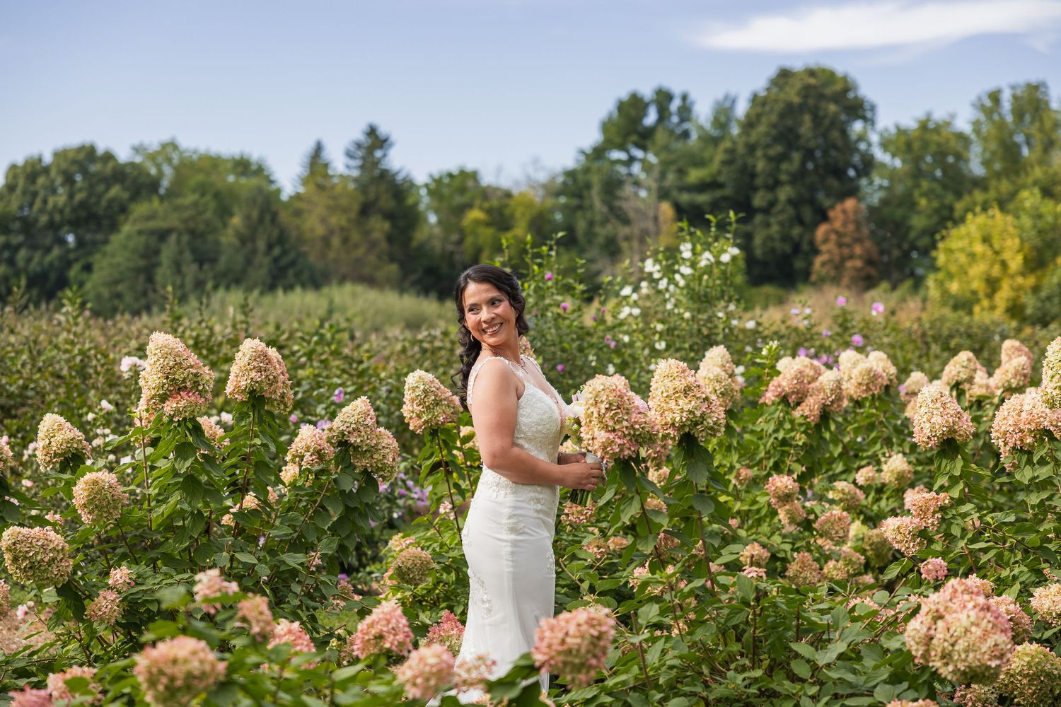 Bride in white dress smiling among pink hydrangeas in a lush garden on her wedding day.