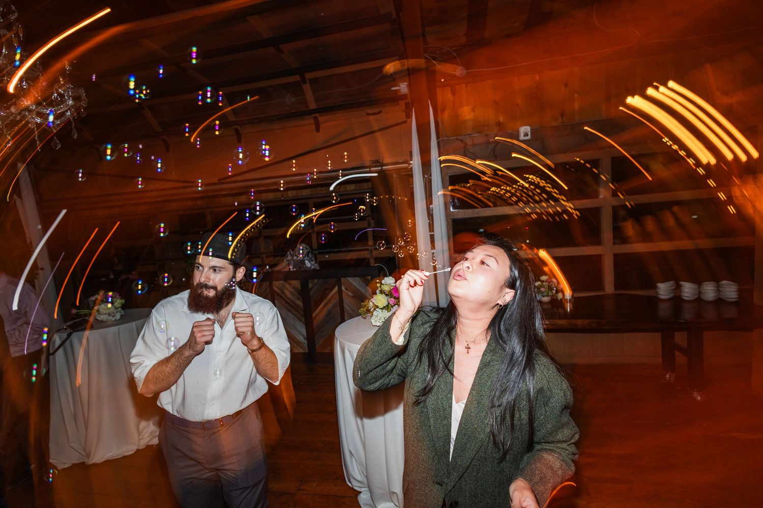 A bearded man and woman blow bubbles at an indoor wedding reception with warm ambient lighting.