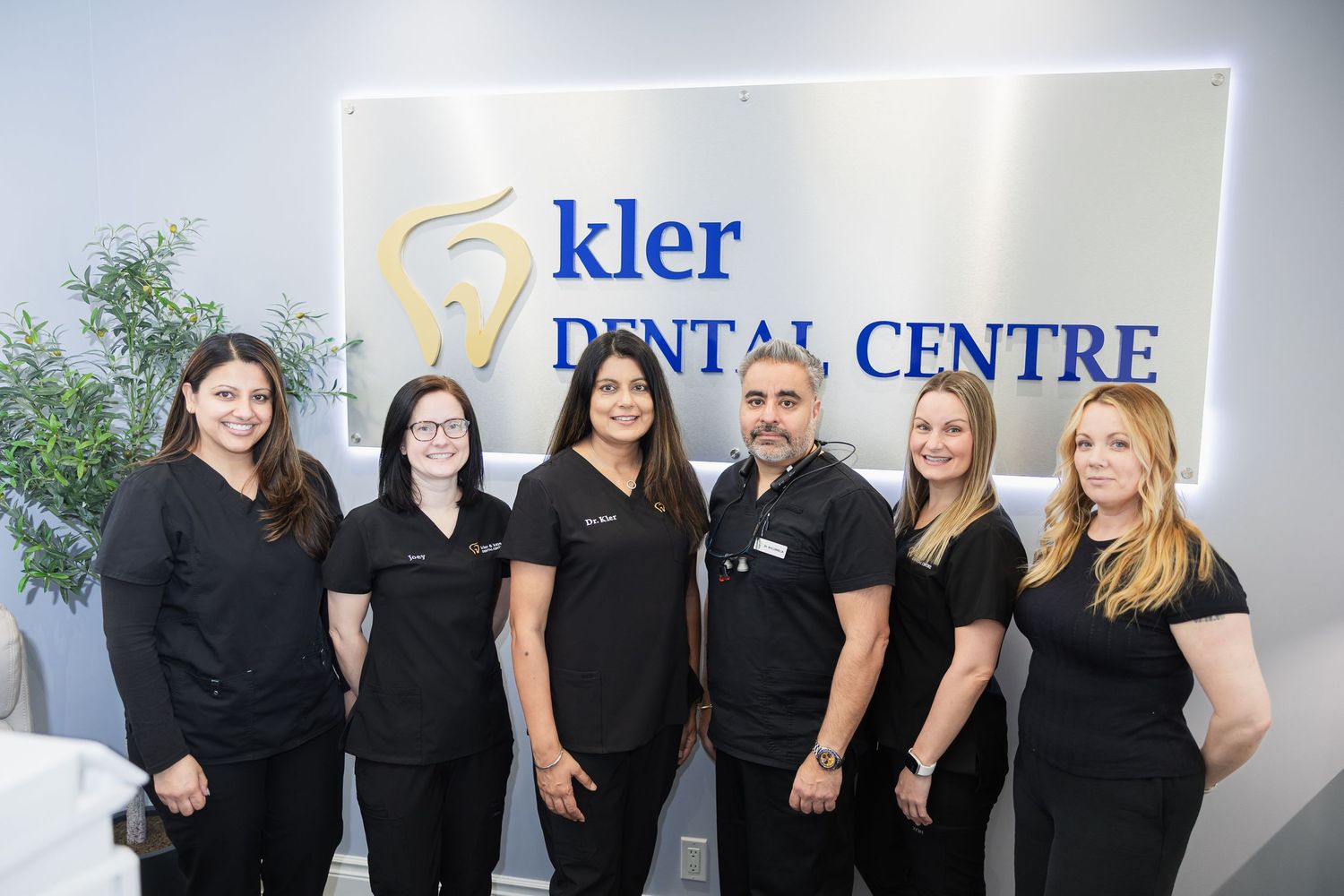 Six dental professionals in black uniforms smiling together at Kler Dental Centre reception area.