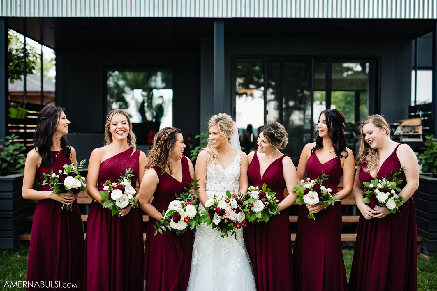 Bridesmaids in burgundy dresses pose together with bouquets at outdoor venue.