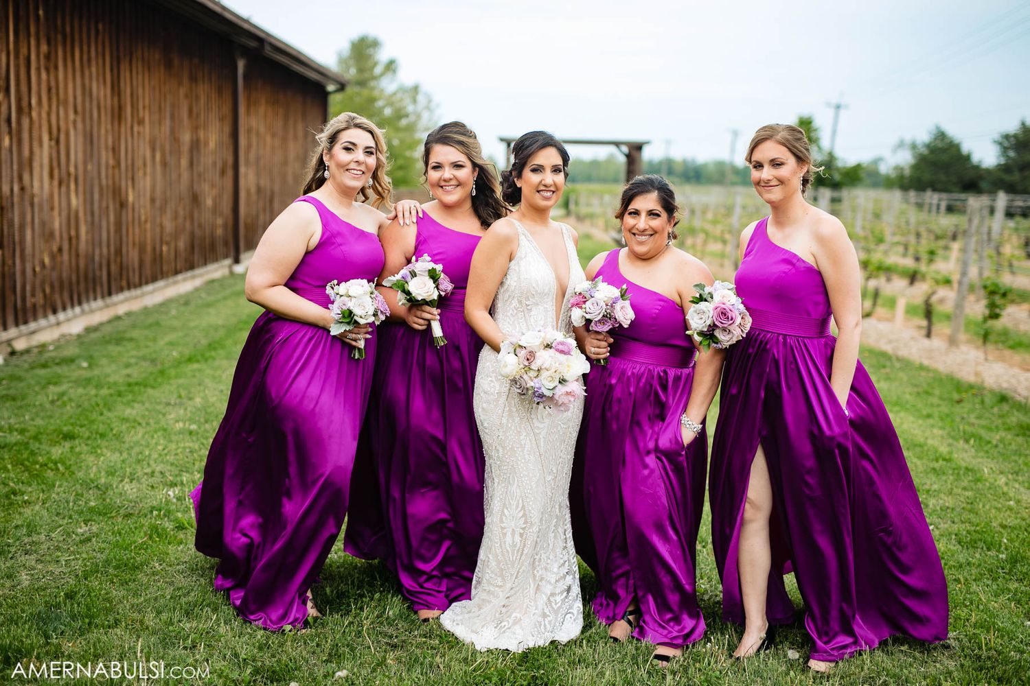 Bridesmaids in vibrant purple dresses pose with bride in white gown outdoors at rustic wedding venue.