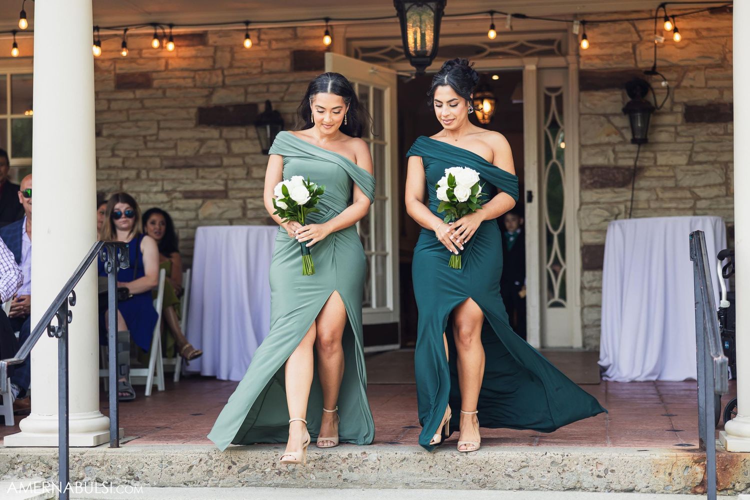Two emerald green gowns with thigh slits and white bouquets on stone steps.