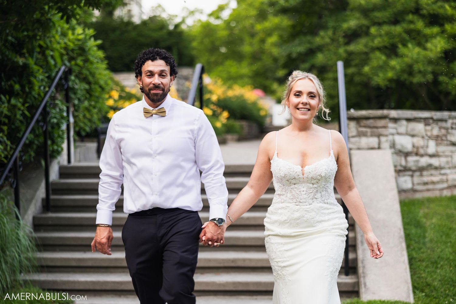 A couple in wedding attire descends stone garden steps while holding hands and smiling.