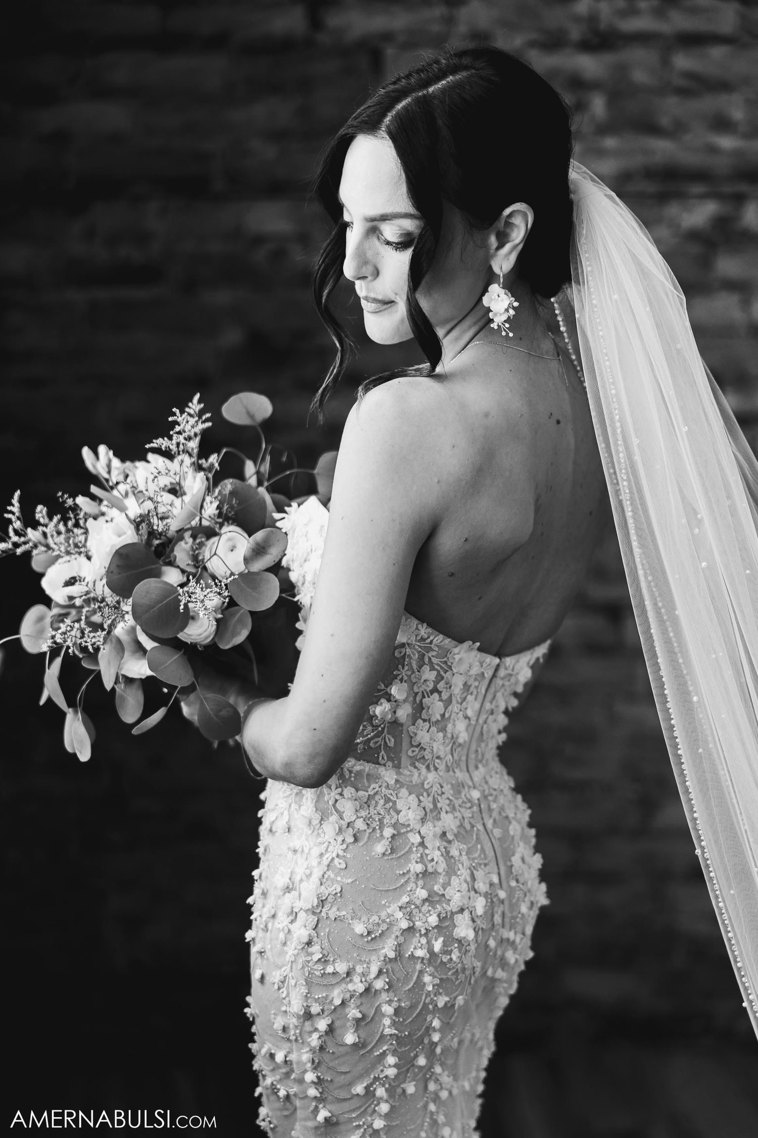 A black and white photo of a bride holding a bouquet against exposed brick walls.