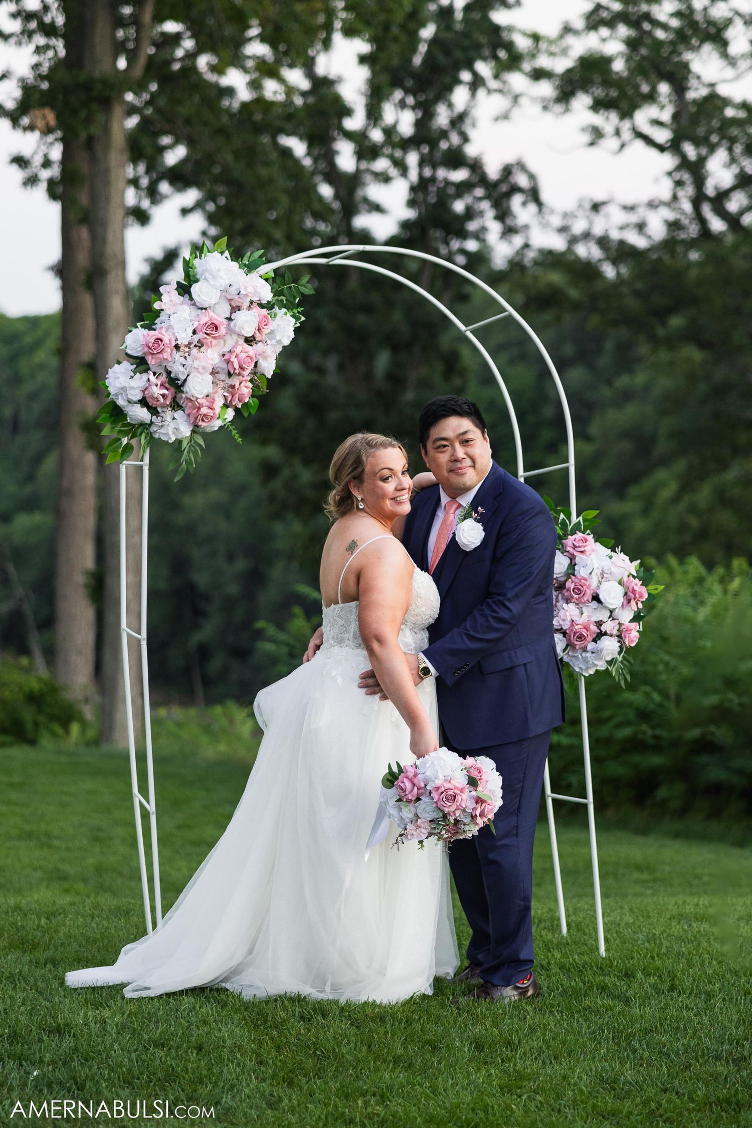 A couple posing under a floral arch decorated with pink and white blooms in a garden setting.