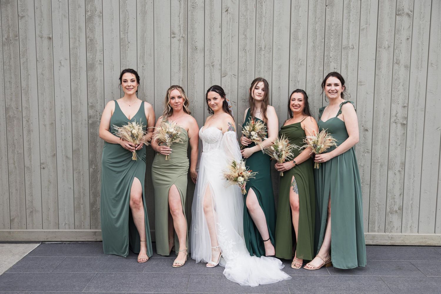Bridesmaids in elegant green dresses pose with bride wearing white gown against grey wall backdrop.