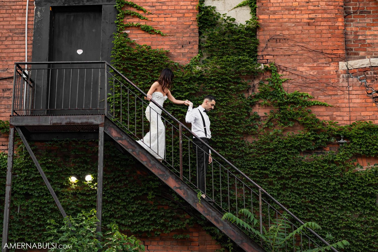 Two figures in white dresses stand on an ivy-covered brick building's fire escape.