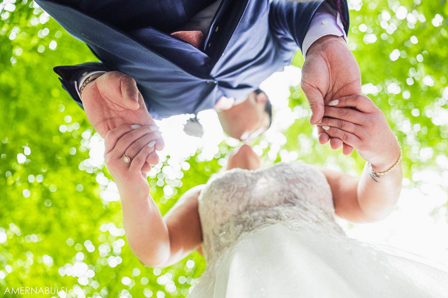 Creative upward angle portrait of newlyweds holding hands against bright green foliage.