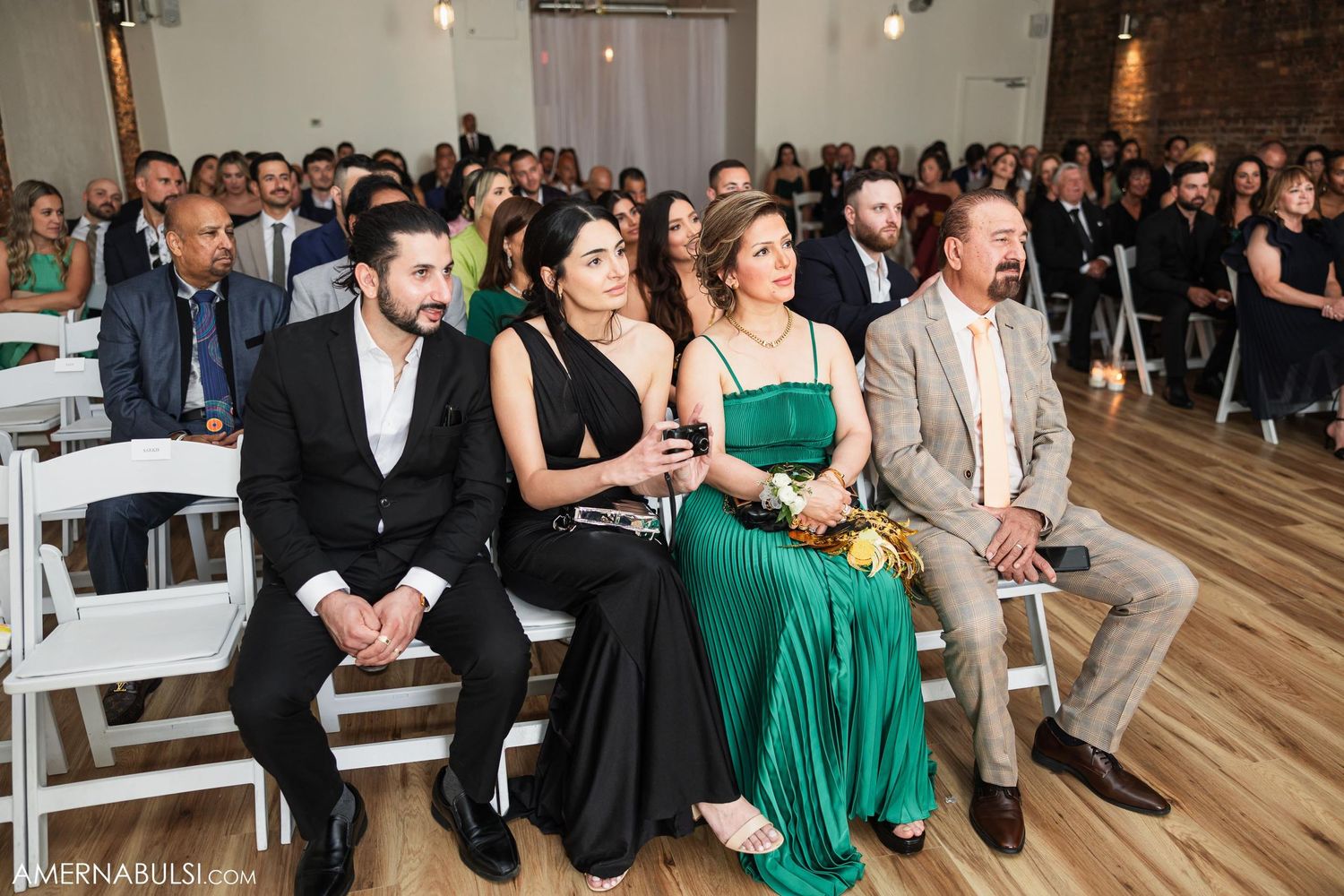 Wedding guests in formal attire seated in white chairs at indoor ceremony venue.