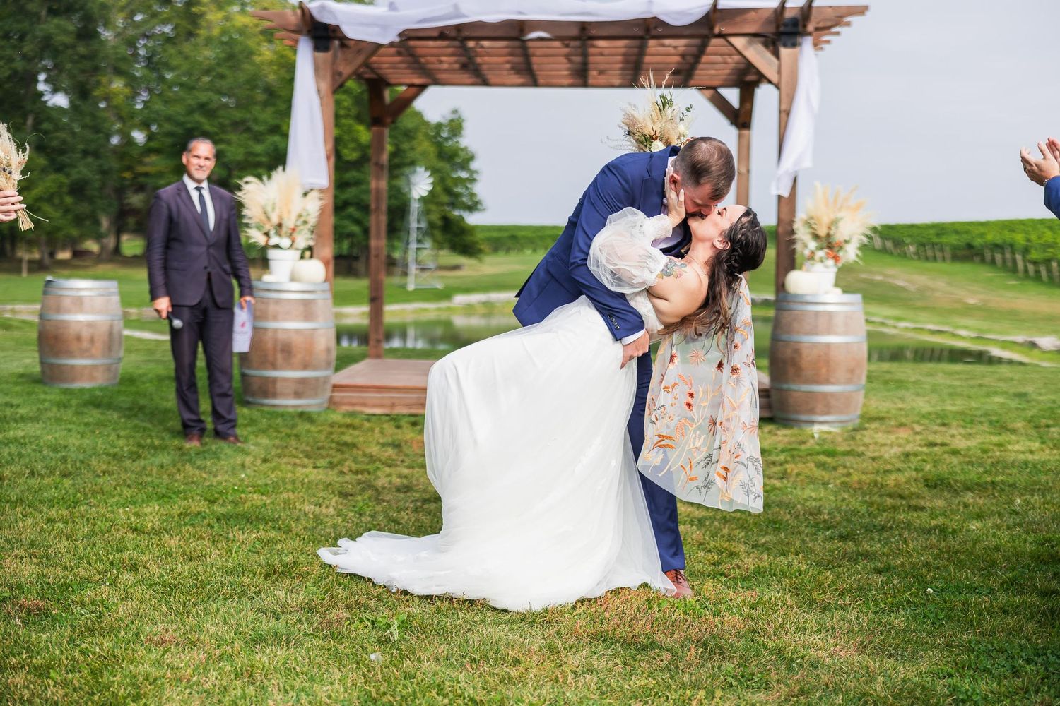 Wedding ceremony moment under wooden pergola with wine barrels in outdoor garden setting.