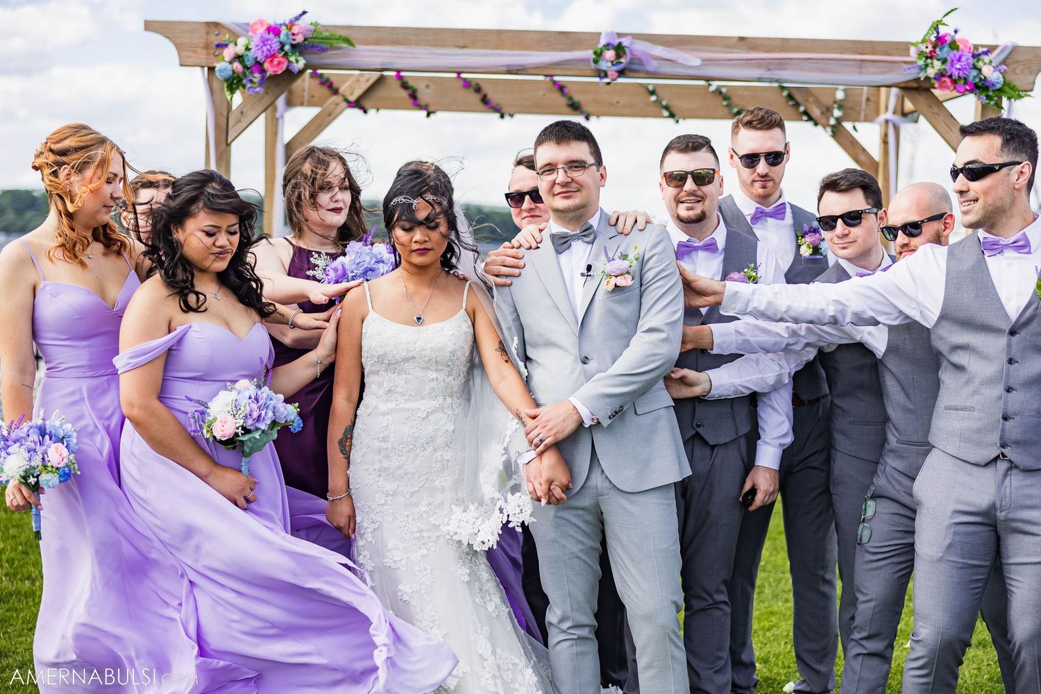 Wedding party in purple bridesmaid dresses and gray suits pose together under wooden arch by waterfront.
