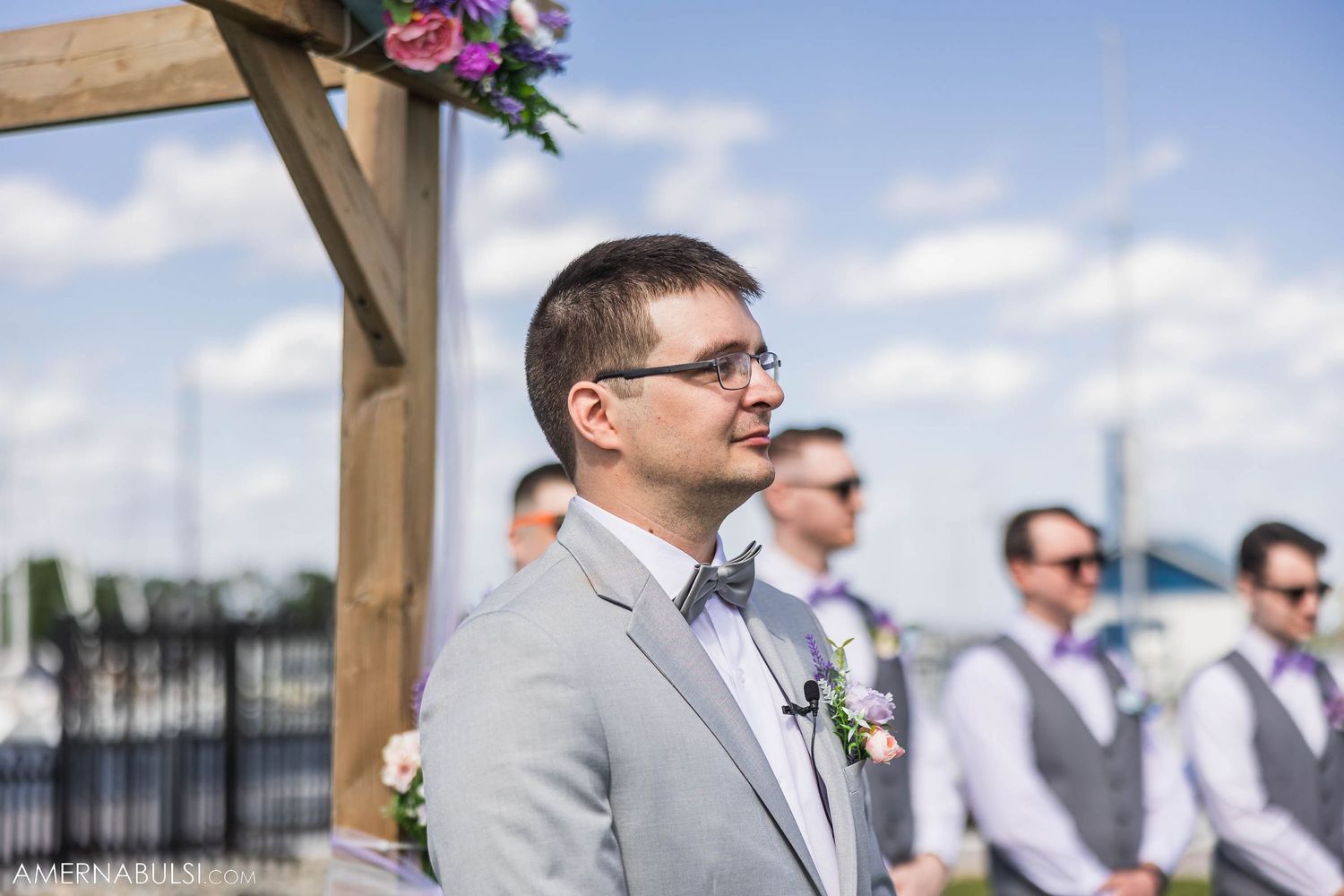 Wedding ceremony setup with wooden arch and groomsmen in light grey suits.