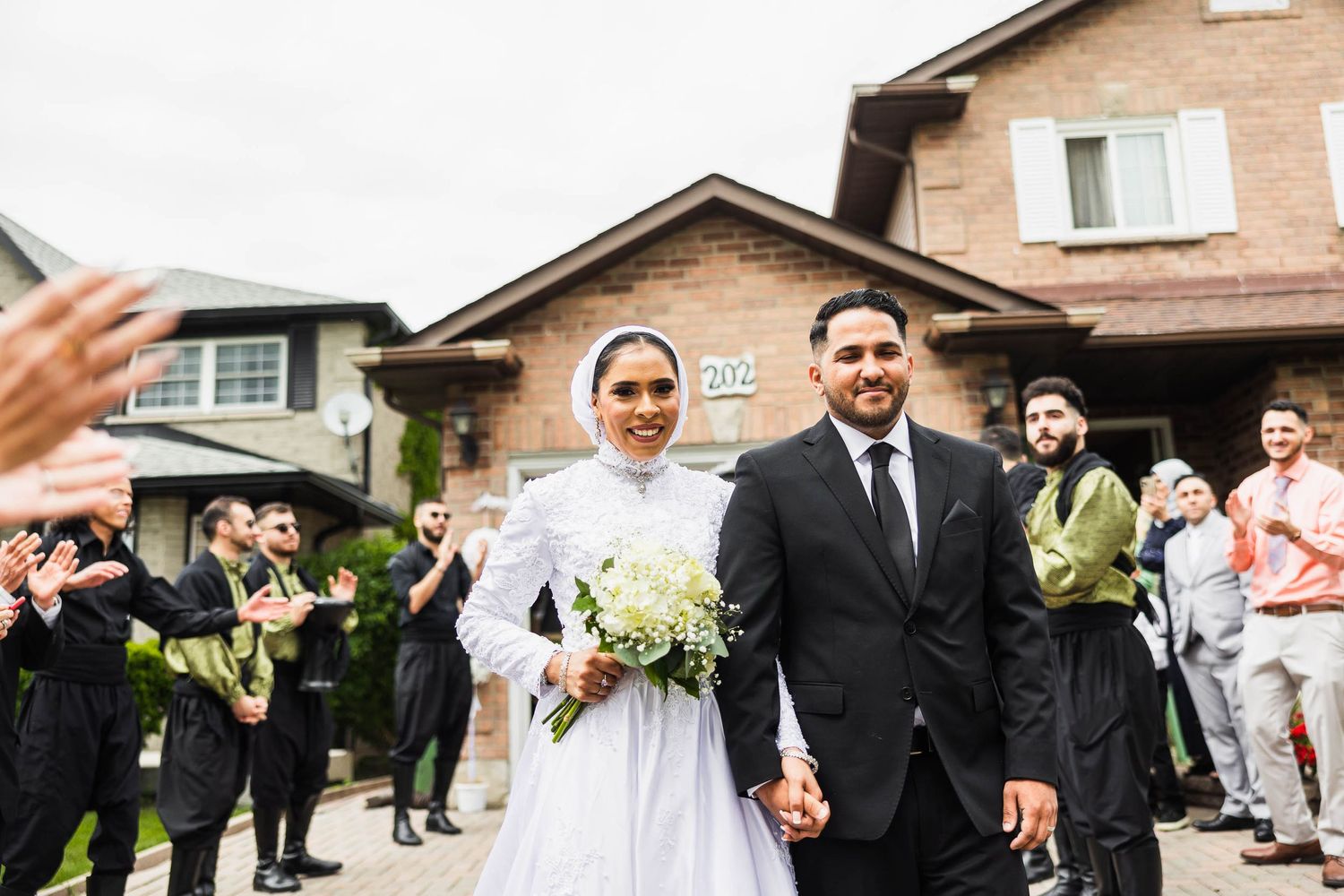 Wedding couple exits ceremony while guests celebrate in residential setting.